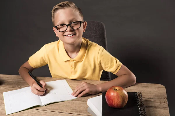 Счастливый школьник в очках смотрит в камеру, делая домашнее задание в учебнике за столом с яблоком и стопкой книг — стоковое фото
