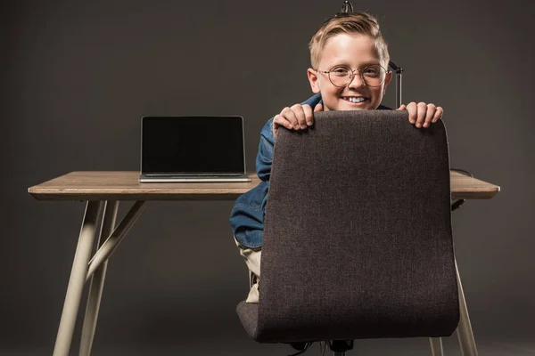 Happy little boy sitting on chair and looking at camera near table with lamp and laptop on grey background — Stock Photo