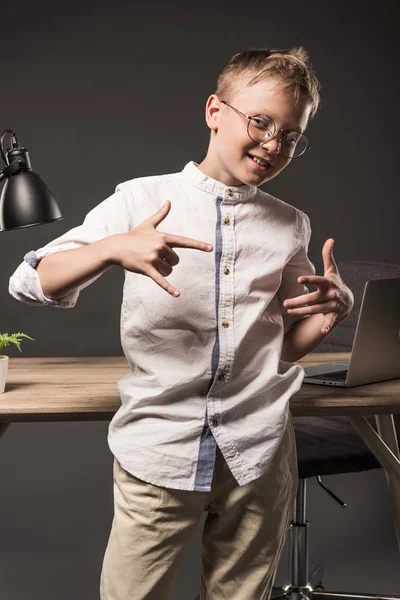 Smiling little boy in eyeglasses gesturing by hands near table with lamp, plant and laptop on grey background — Stock Photo