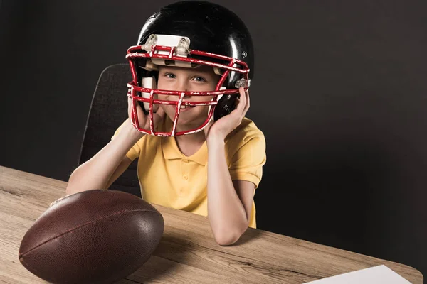 Kleiner lächelnder Junge mit Schutzhelm am Tisch mit American Football Ball auf grauem Hintergrund — Stockfoto