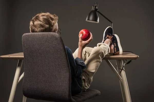 Vista trasera del niño descansando con las piernas sobre la mesa y sosteniendo la manzana sobre fondo gris - foto de stock