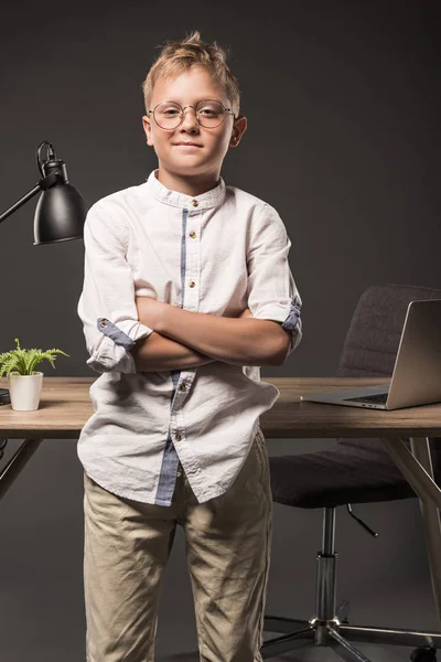 Маленький мальчик в очках со скрещенными руками, стоящий рядом со столом с растением, ноутбуком и лампой на сером фоне — стоковое фото