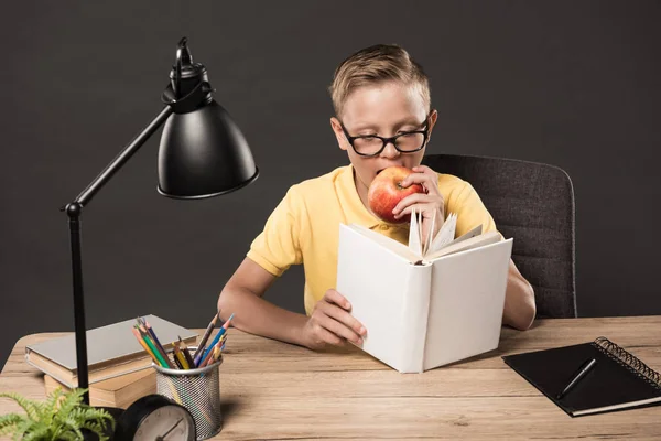 Colegial serio en anteojos leyendo libro y comiendo manzana en la mesa con lápices de color, reloj, lámpara, pila de libros y libro de texto sobre fondo gris - foto de stock