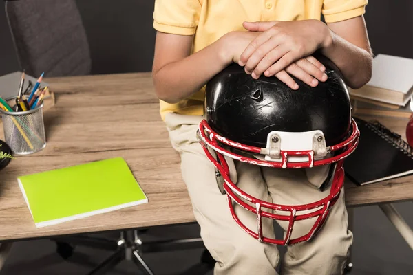 Обрезанное изображение школьника, держащего американский футбольный шлем и сидящего на столе с книгами, цветными карандашами и учебниками на сером фоне — стоковое фото