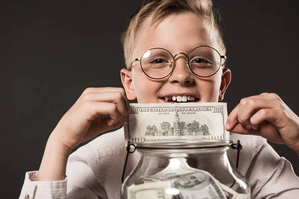Portrait of smiling little boy in eyeglasses showing dollar banknote over jar full of cash money isolated on grey background — Stock Photo