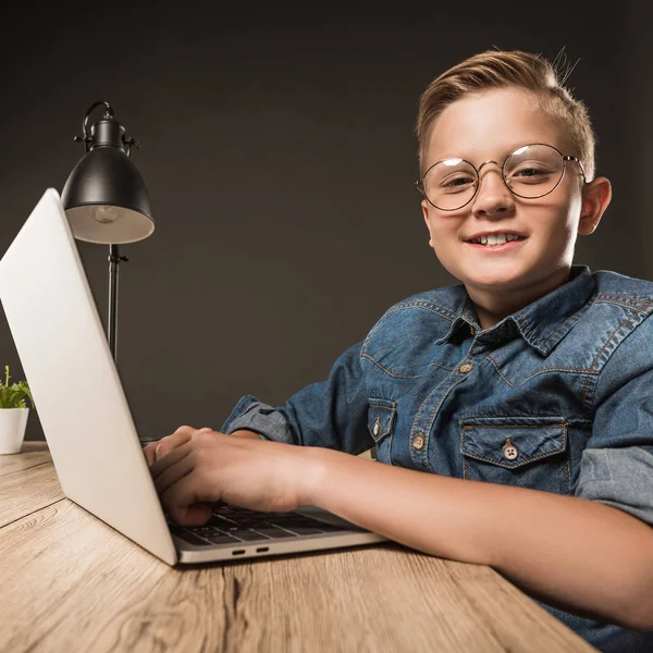 Smiling little boy in eyeglasses using laptop at table with lamp and plant on grey background — Stock Photo