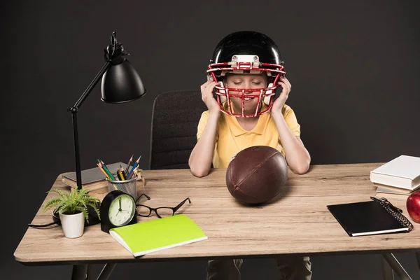 Школьник надевает американский футбольный шлем и сидит с ногами на столе с мячом, книгами, растением, лампой, цветными карандашами, яблоком, часами и учебниками на сером фоне — стоковое фото