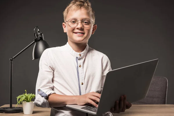 Niño feliz en gafas con portátil y mirando a la cámara cerca de la mesa con lámpara y la planta sobre fondo gris - foto de stock