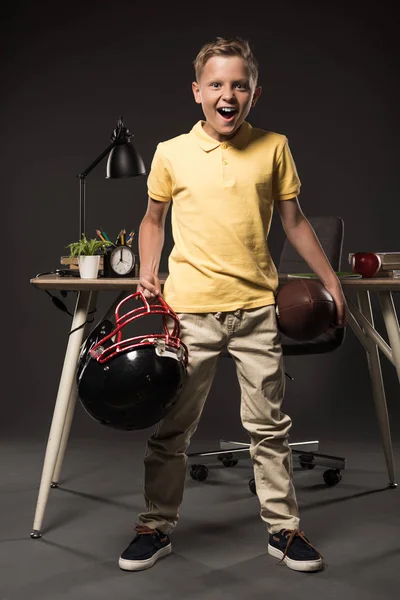 Colegial gritando emocionado sosteniendo casco de fútbol americano con pelota y de pie cerca de la mesa con libros, planta, lámpara, lápices de color, manzana, reloj y libro de texto sobre fondo gris - foto de stock