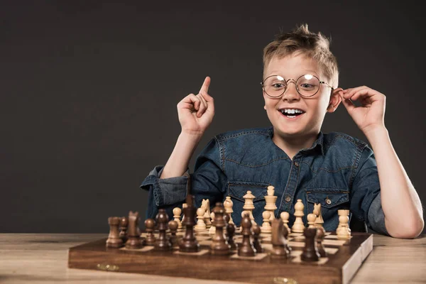 Niño feliz en anteojos haciendo gesto de idea con el dedo en la mesa con tablero de ajedrez aislado sobre fondo gris - foto de stock