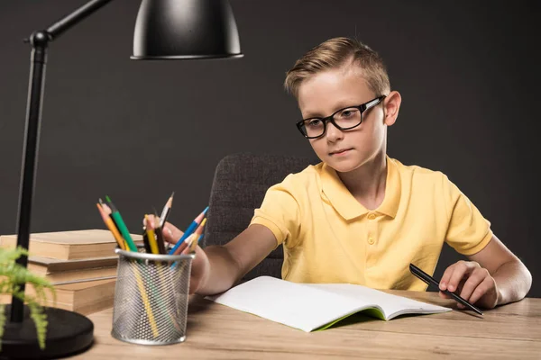 Serious schoolboy in eyeglasses doing homework at table with colour pencils, books, lamp and textbook on grey background — Stock Photo