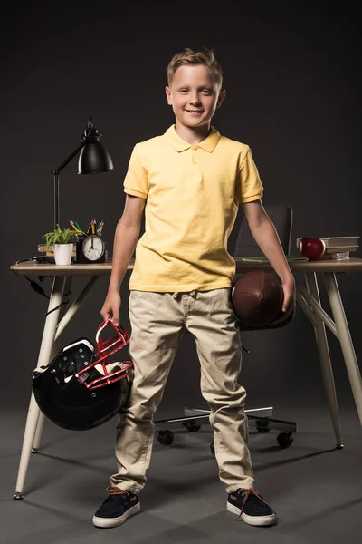 Adorable colegial sosteniendo casco de fútbol americano con pelota y de pie cerca de la mesa con libros, planta, lámpara, lápices de color, manzana, reloj y libro de texto sobre fondo gris - foto de stock