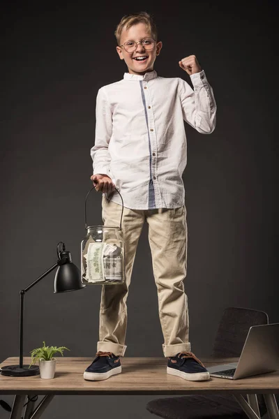 Happy little boy in eyeglasses holding jar full of dollar banknotes and gesturing by hand while standing on table with laptop, plant and lamp on grey background — Stock Photo