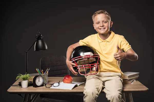 Школьник держит американский футбольный шлем, делает большой палец вверх жестом и сидит на столе с книгами, очками, растением, лампой, цветными карандашами, яблоком, часами и учебником на сером фоне — стоковое фото