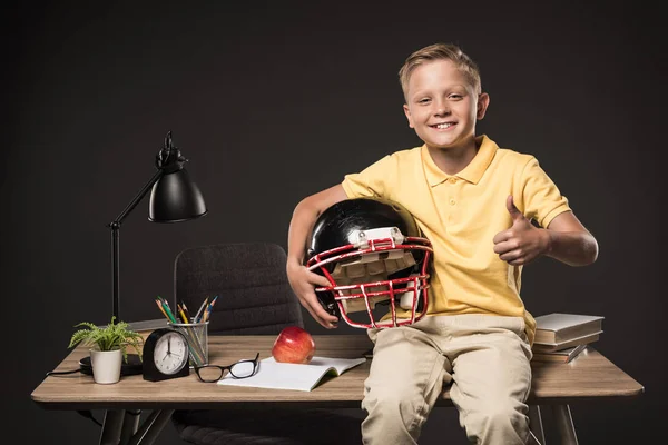 Colegial sosteniendo casco de fútbol americano, haciendo gesto de pulgar hacia arriba y sentado en la mesa con libros, anteojos, planta, lámpara, lápices de color, manzana, reloj y libro de texto sobre fondo gris - foto de stock