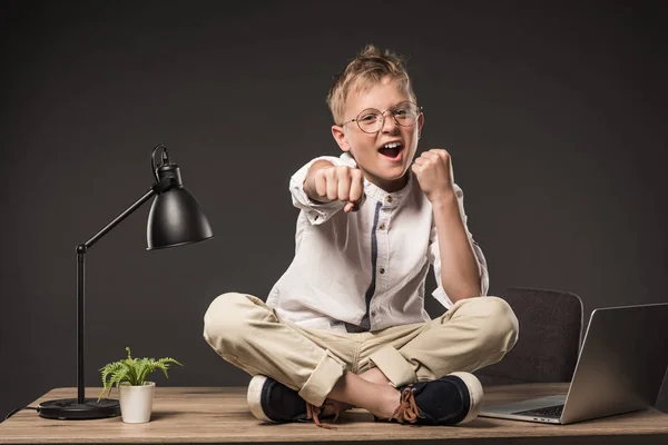 Excited little boy in eyeglasses fighting by fists while sitting on table with lamp, plant and laptop on grey background — Stock Photo