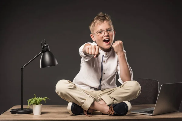 Excité petit garçon dans les lunettes de combat par les poings tout en étant assis sur la table avec lampe, plante et ordinateur portable sur fond gris — Photo de stock