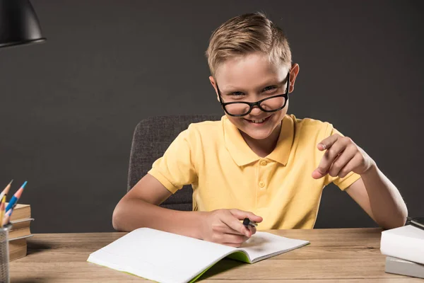 Smiling schoolboy in eyeglass pointing by finger and doing homework at table with books, colour pencils and textbook on grey background — Stock Photo