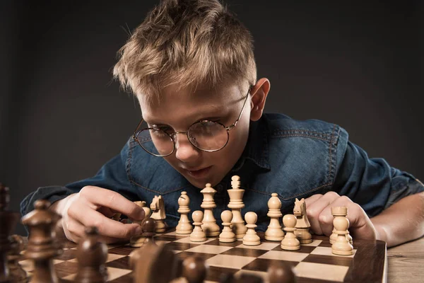 Focused little boy in eyeglasses playing chess at table isolated on grey background — Stock Photo