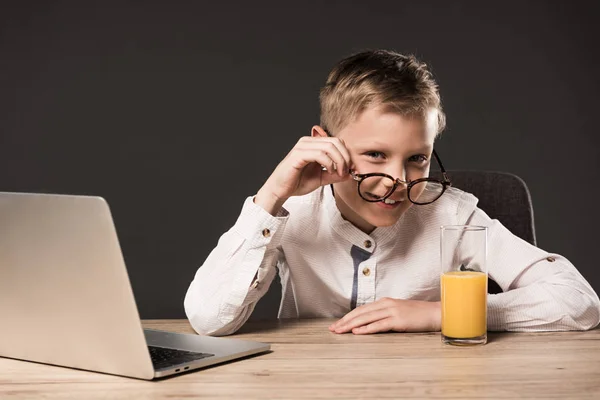 Smiling little boy putting on eyeglasses at table with laptop and glass of juice on grey background — Stock Photo