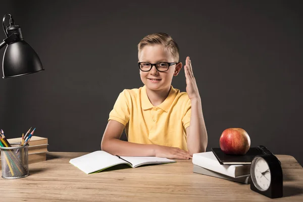 Улыбающийся школьник с поднятой рукой сидит за столом с часами, лампой, цветными карандашами, книгами, учебниками и яблоками на сером фоне — стоковое фото