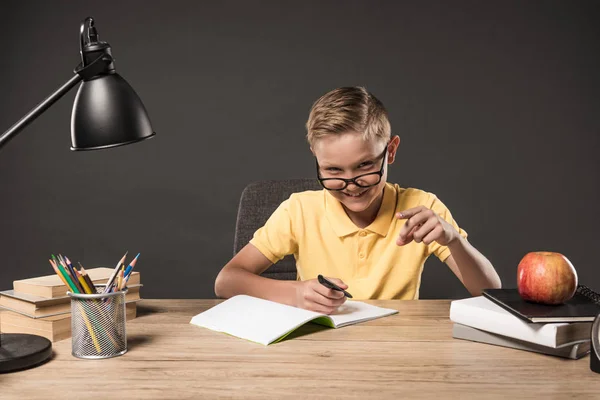 Colegial feliz en gafas apuntando con el dedo y haciendo los deberes en la mesa con lámpara, libros, lápices de color y libro de texto sobre fondo gris - foto de stock