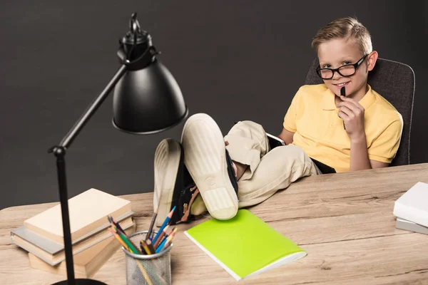 Colegial sonriente en gafas haciendo deberes con piernas en la mesa con lámpara, libros, lápices de color y libro de texto sobre fondo gris - foto de stock