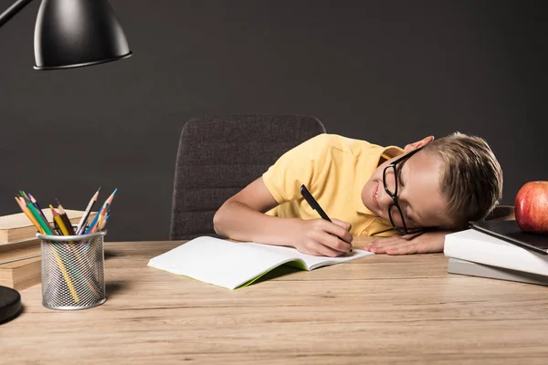 Schoolboy in eyeglass laying on table and doing homework at table with lamp, books, colour pencils and textbook on grey background — Stock Photo