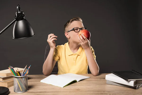 Школьник в очках ест яблоко и делает домашнее задание за столом с лампой, книгами, цветными карандашами и учебником на сером фоне — стоковое фото