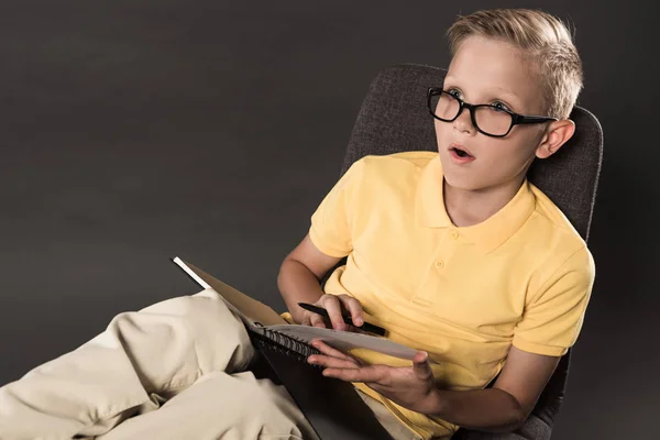 Шокированный школьник в очках сидит на стуле и делает домашнее задание с учебником на сером фоне — стоковое фото