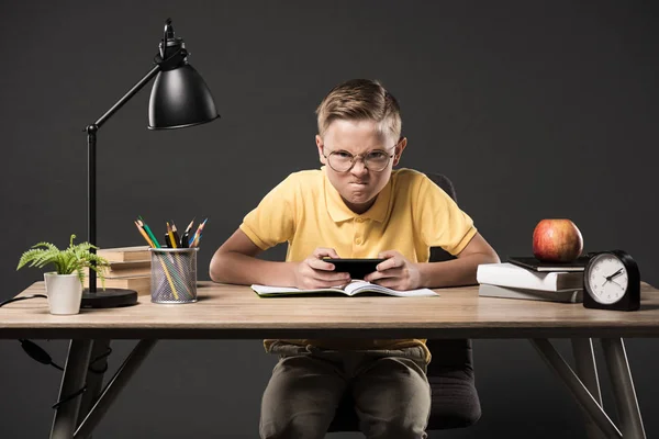 Angry schoolboy in eyeglasses using smartphone and sitting at table with books, plant, clock, plant, colour pencils, lamp, textbooks on grey background — Stock Photo
