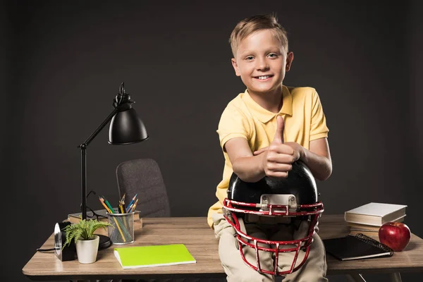 Feliz colegial sosteniendo casco de fútbol americano, haciendo gesto pulgar hacia arriba y sentado en la mesa con libros, planta, lámpara, lápices de color, manzana, reloj y libro de texto sobre fondo gris - foto de stock