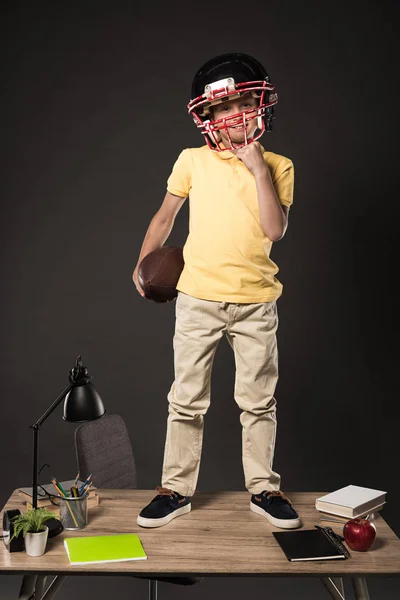 Школьник в американском футбольном шлеме держит мяч и стоит на столе с книгами, растением, лампой, цветными карандашами, яблоком, часами и учебником на сером фоне — стоковое фото