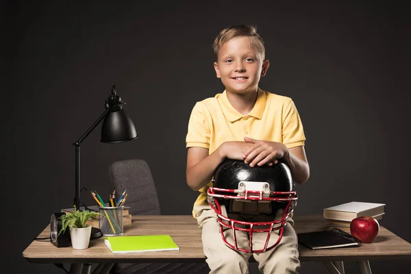 Усміхнений школяр тримає американський футбольний шолом і сидить на столі з книгами, рослинами, лампами, кольоровими олівцями, яблуком, годинником і підручником на сірому фоні — стокове фото