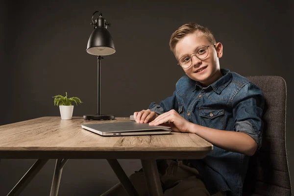 Smiling little boy in eyeglasses opening laptop at table with plant and lamp on grey background — Stock Photo