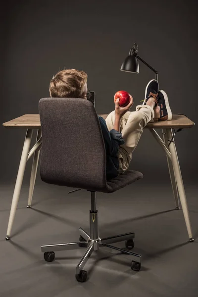 Вид сзади маленького мальчика, держащего яблоко в руке, когда он отдыхает с ногами на столе с лампой и ноутбуком на сером фоне — стоковое фото