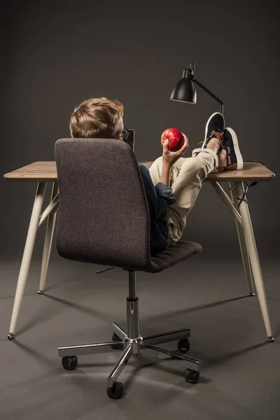 Rear view of little boy holding apple in hand while resting with legs on table with lamp and laptop on grey background — Stock Photo