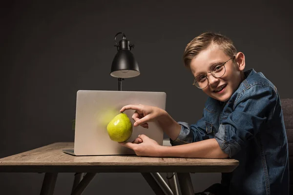 Lächelnder kleiner Junge in Brille mit Birne vor Laptop am Tisch mit Laptop auf grauem Hintergrund — Stockfoto