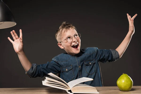 Возбужденный школьник в очках жестикулирует руками, читая книгу за столом с лампой и грушей на сером фоне — стоковое фото