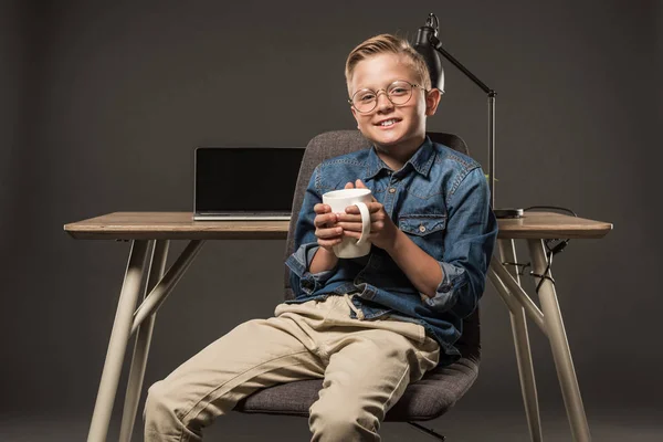 Smiling little boy in eyeglasses sitting on chair with coffee cup near table with laptop and lamp on grey background — Stock Photo