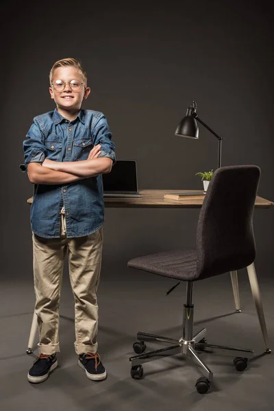 Niño pequeño en gafas de pie con las manos cruzadas cerca de la mesa con el ordenador portátil, libro y lámpara - foto de stock
