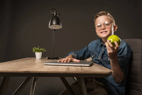 Heureux petit garçon dans les lunettes tenant poire à table avec ordinateur portable, plante et lampe sur fond gris — Photo de stock