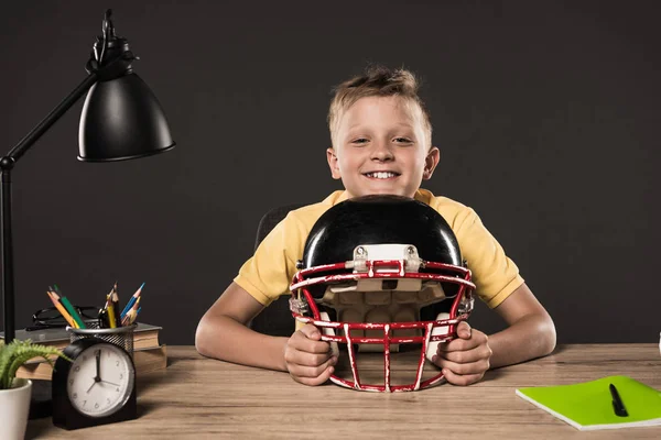 Heureux écolier avec casque de football américain assis avec à la table avec des livres, plante, lampe, crayons de couleur et manuel sur fond gris — Photo de stock