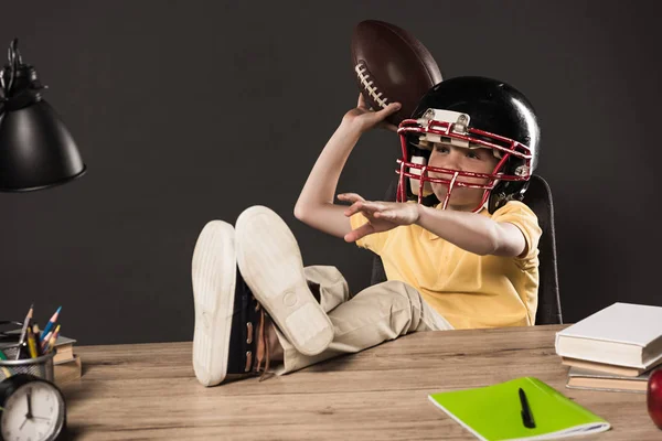 Estudante no capacete de futebol americano jogando bola e sentado com as pernas na mesa com livros, planta, lâmpada, lápis de cor, maçã, relógio e livro didático sobre fundo cinza — Fotografia de Stock