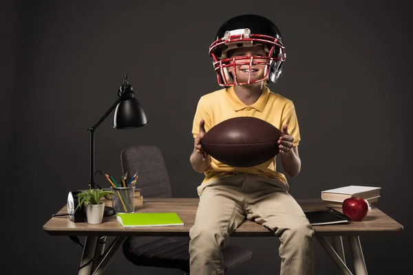 Lächelnder Schuljunge mit American-Football-Helm, Ball in der Hand und auf Tisch sitzend mit Büchern, Pflanze, Lampe, Farbstiften, Apfel, Uhr und Lehrbuch auf grauem Hintergrund — Stockfoto