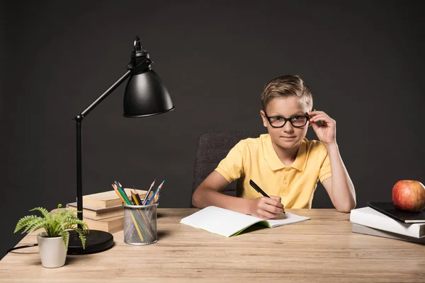 Schoolboy in eyeglasses doing homework at table with books, plant, lamp, colour pencils, apple, clock and textbook on grey background — Stock Photo