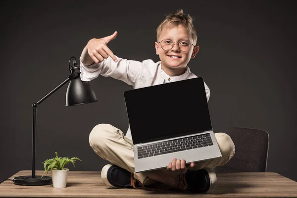 Усміхнений маленький хлопчик в окулярах сидить на столі і вказує на ноутбук з порожнім екраном на сірому фоні — стокове фото