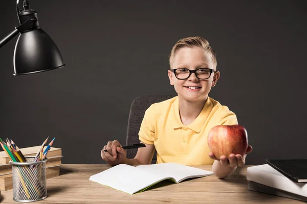 Scolaro in occhiali che tiene la mela e fa i compiti a tavola con libri, lampada, matite a colori e libro di testo su sfondo grigio — Foto stock