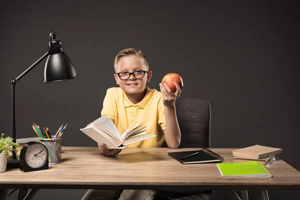 Усміхнений школяр в окулярах тримає яблуко і читає книгу за столом з книгами, рослинами, лампами, кольоровими олівцями, годинником і підручником на сірому фоні — стокове фото