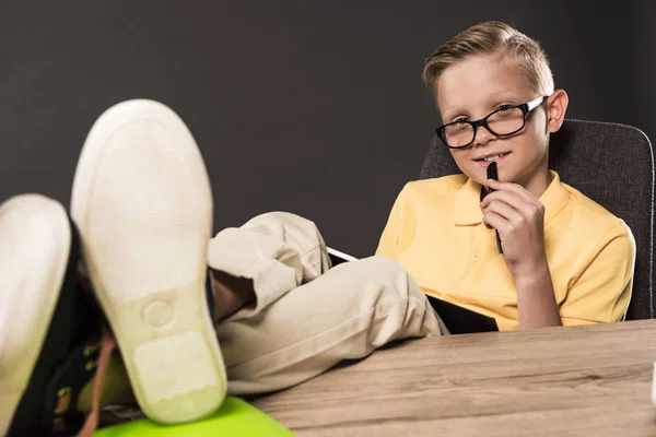 Schoolboy in eyeglasses with textbook doing homework with legs on table on grey background — Stock Photo